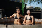 Two women in black merino wool triangle bikini tops sitting in an outdoor cold plunge tub
