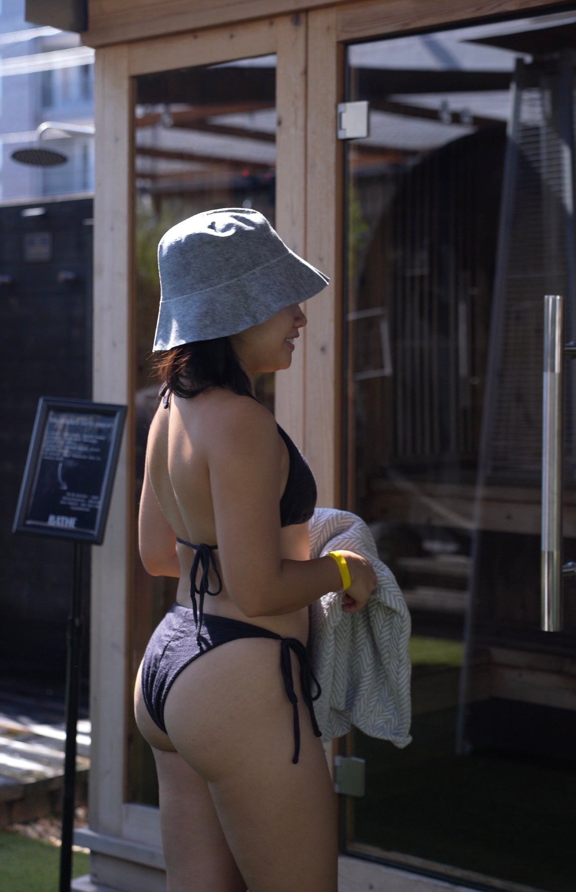 Back view of a woman in a black merino wool bikini and wool sauna hat, standing outside a sauna