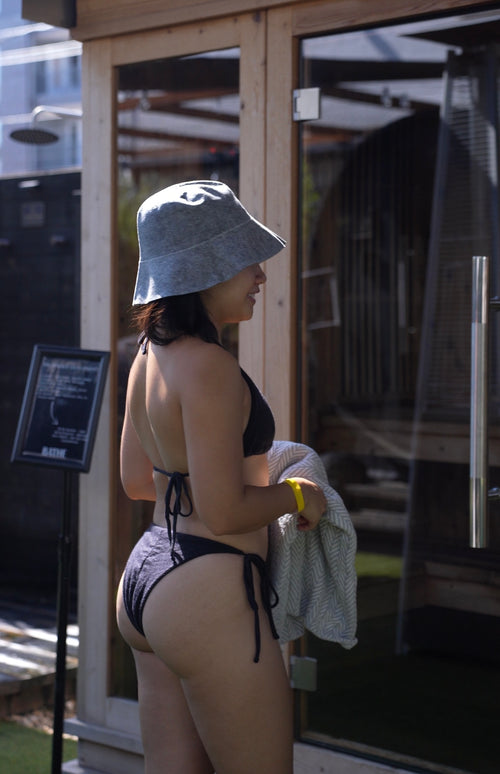 Back view of a woman in a black merino wool bikini and wool sauna hat, standing outside a sauna